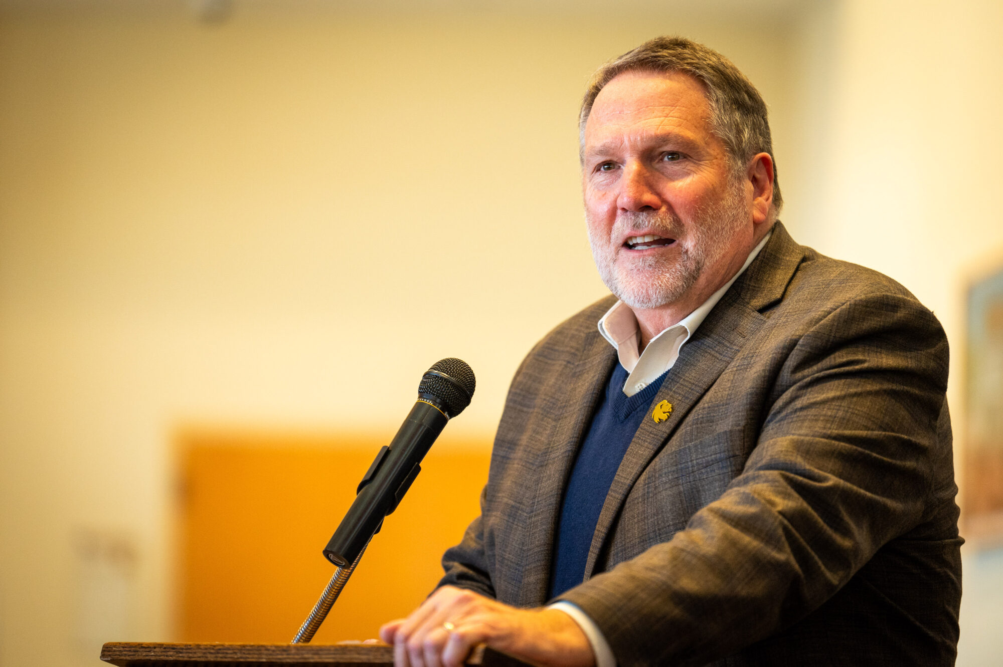 A person stands behind a podium with his left hand on the podium. He speaks into the microphone. He is wearing a suit jacket with a lion head on the lapel, and he smiles as he speaks.