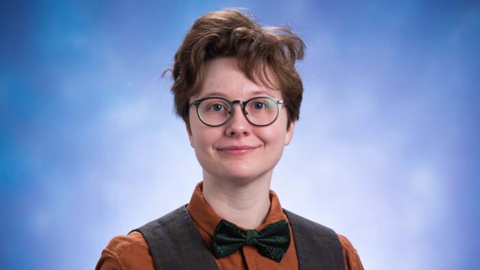Person smiles for professional headshot photo, wearing a black bowtie, black vest, rust-colored shirt and glasses.