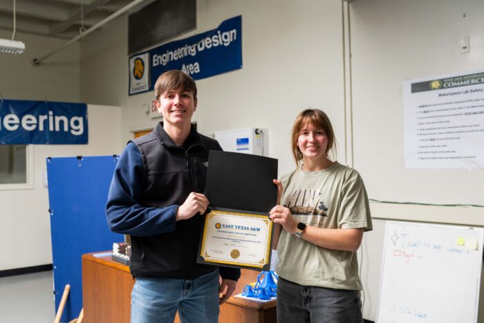 Two people smile at the camera, holding a certificate between them. An engineering lab is shown behind them.