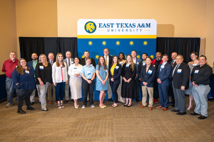 A large group of people face the camera and smile. In the background, a blue backdrop with yellow lion heads is visible, along with a white sign above the backdrop with the words "East Texas A&M."