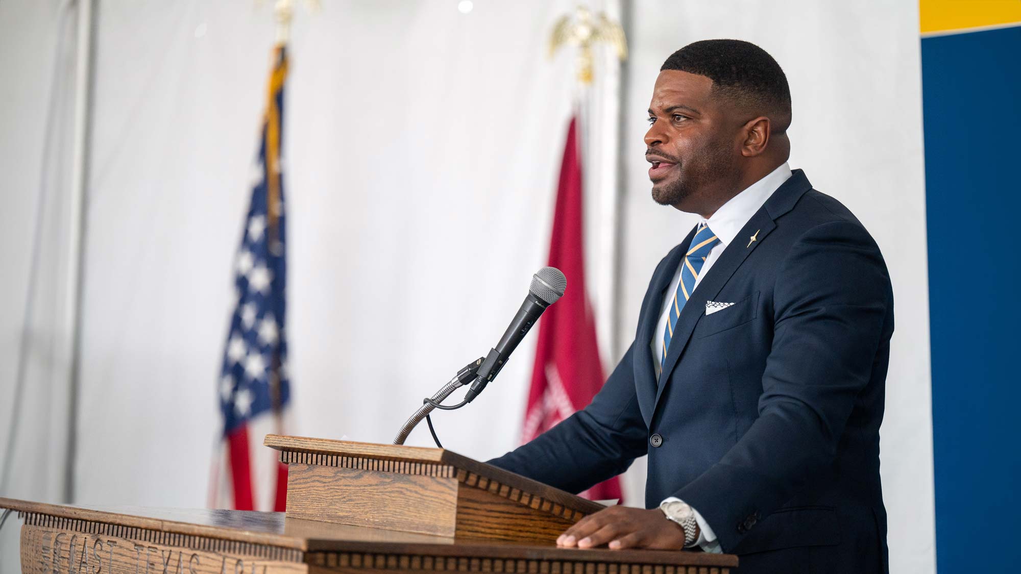 Speaker stands behind microphone with hands on podium. American flag and A&M System flag are shown in the background. Speaker wears a suit and tie.