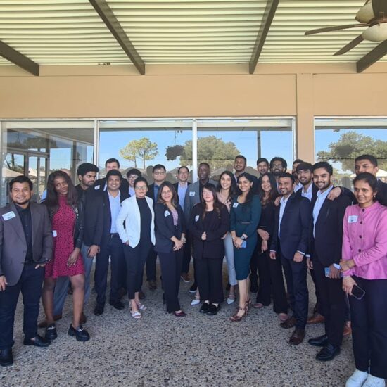A group of professionals, dressed in business attire, pose together under a pergola outside a building. Trees and a clear sky are visible reflecting off the windows in the background.