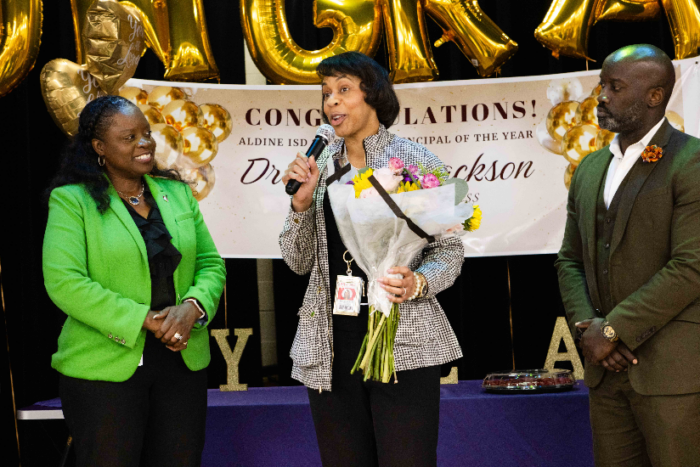 Three people are standing on stage. The person in the middle is holding a microphone and flowers to accept an award.