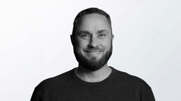 A black and white photograph of a man with a beard and a black tshirt, smiling at the camera.
