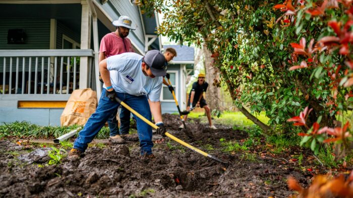 Students dig in the dirt with shovels in hand. A blue wood frame house is shown in the background.