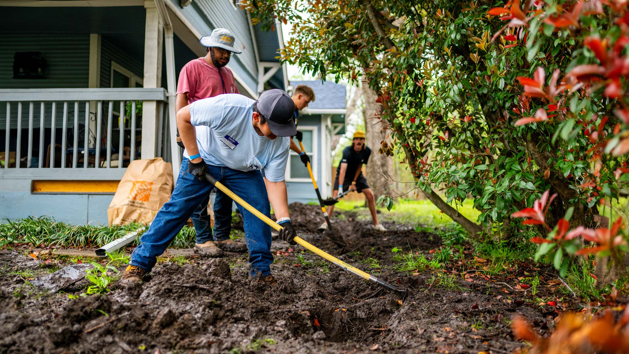 Students dig in the dirt with shovels in hand. A blue wood frame house is shown in the background.