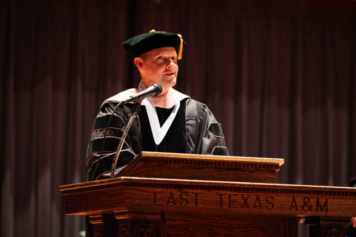 A college faculty member in graduation regalia speaks at a large wooden podium engraved with the name of East Texas A&M University.