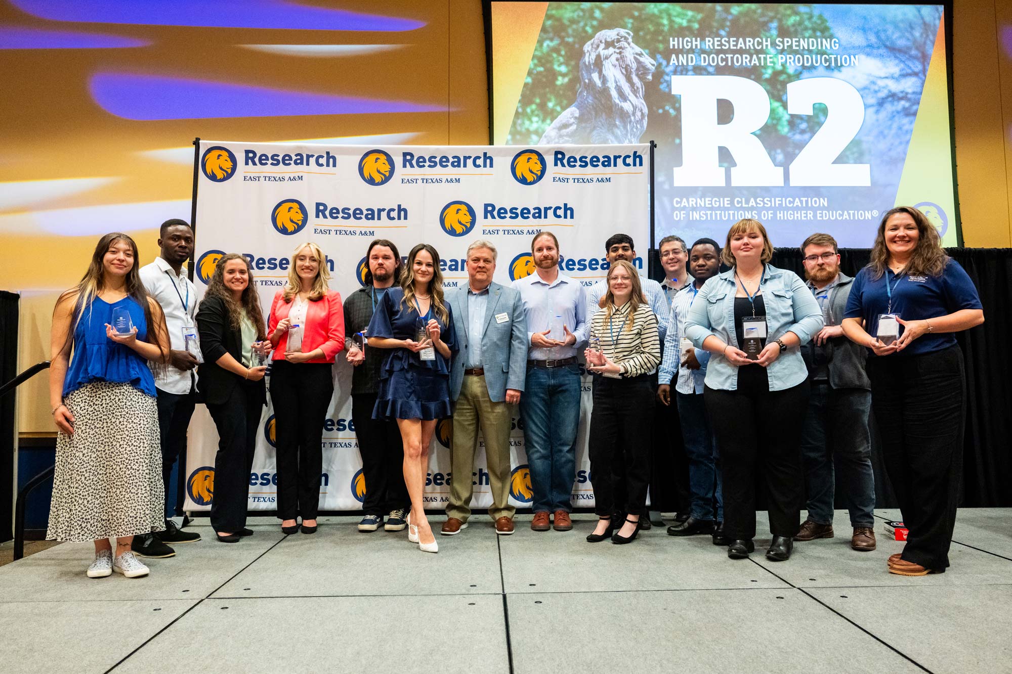 A group of people stands in front of a backdrop that says "Research" and "East Texas A&M" with a gold lion head logo. A large screen is also hanging above the group with "R2" displayed on it.