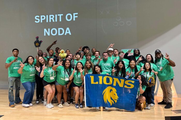 A group of 20+ students wears green shirts with black shorts and hold a flag in front of them with the word "Lion" on it with the university's Lion head logo.