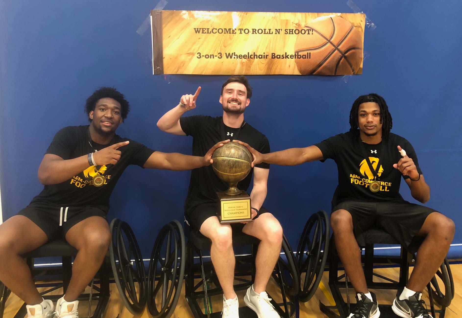 Three basketball players sitting in wheelchairs, one with a trophy in his lap.