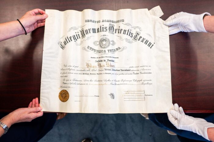 An off-white diploma is written in Latin. White-gloved hands hold the diploma on the right and ungloved hands hold the diploma on the left.