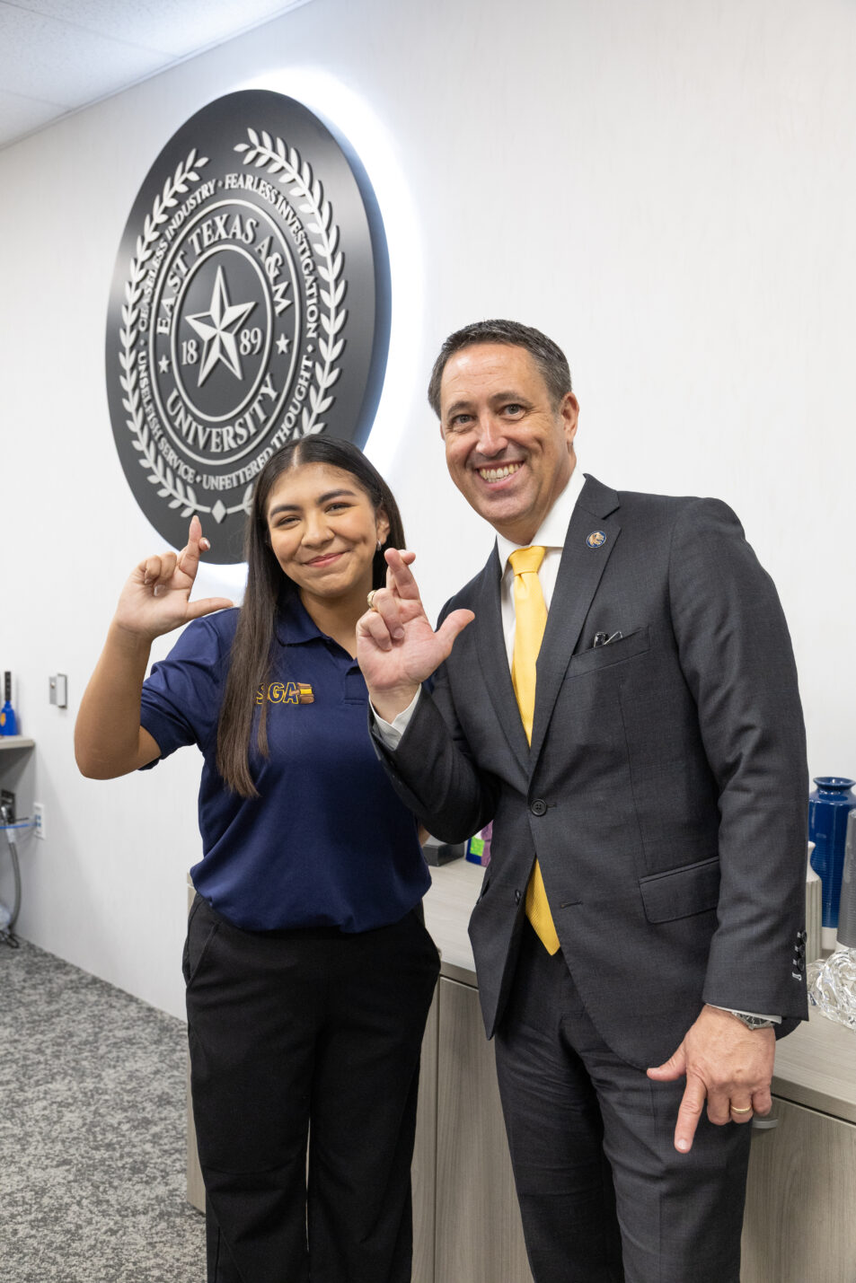 Two people face the camera and smile, making an "L" shape with their right hands and fingers.