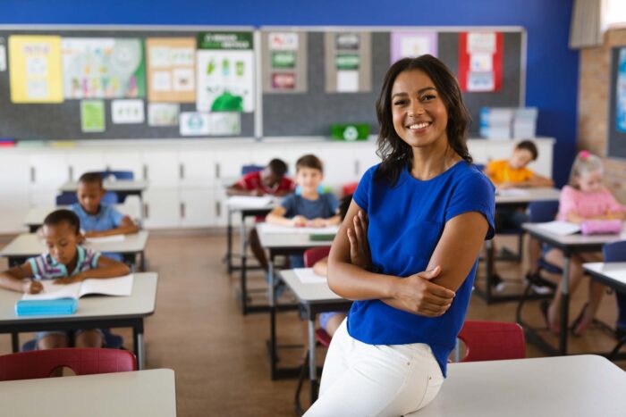Teacher stands in front of classroom, facing camera, with arms folded. She is smiling and her students are sitting in desks behind her.