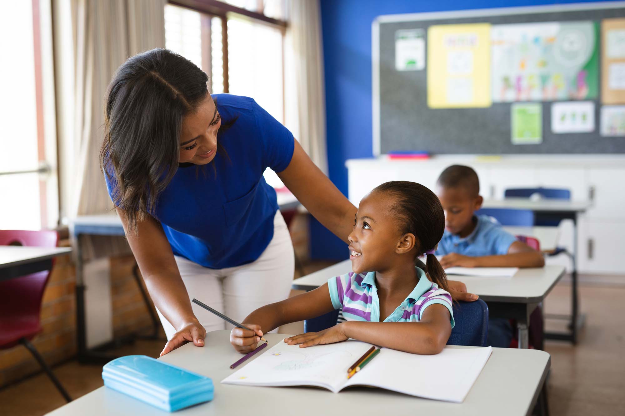Teacher leans forward and faces student at student's desk. Student has a pencil in hand and is working from a workbook.