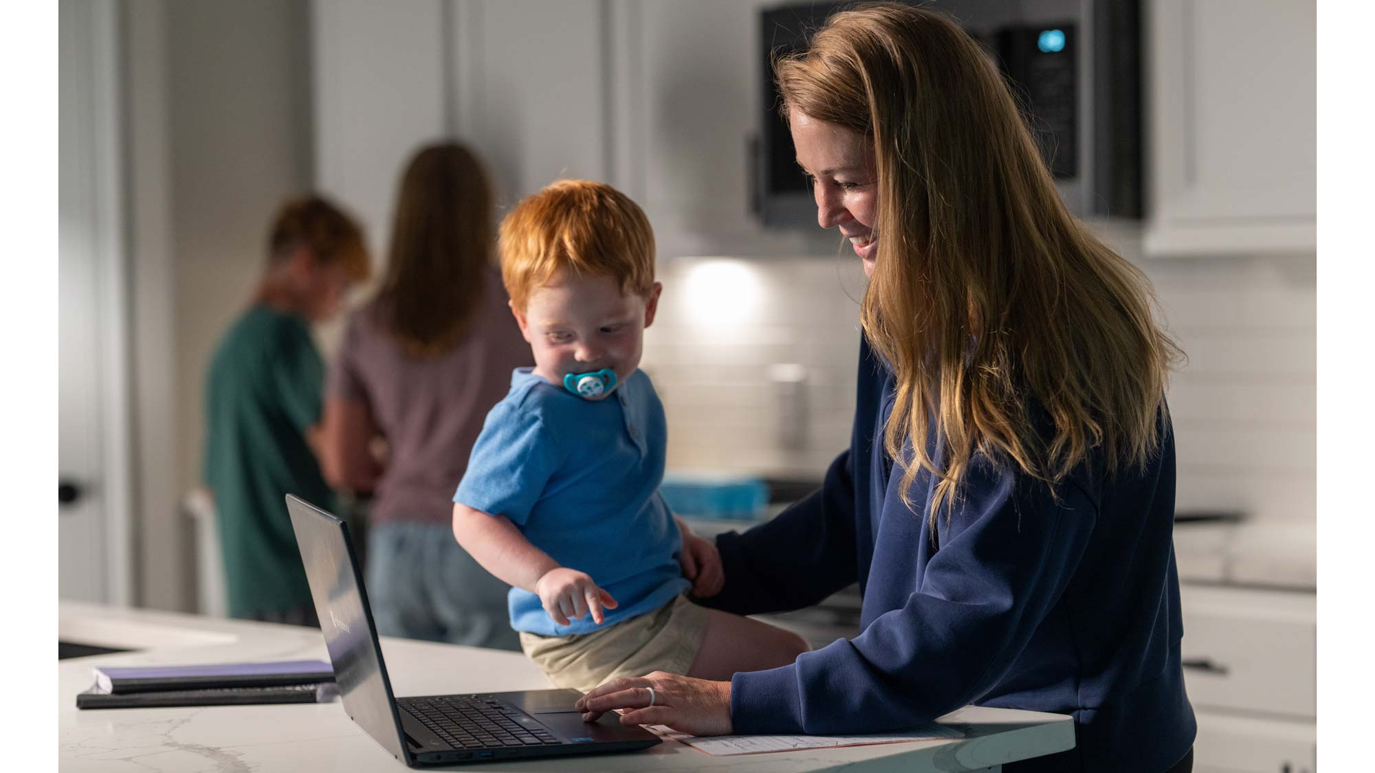 Person is smiling and looking at laptop. A toddler is sitting on the kitchen counter beside her laptop. He is looking at the computer and smiling.