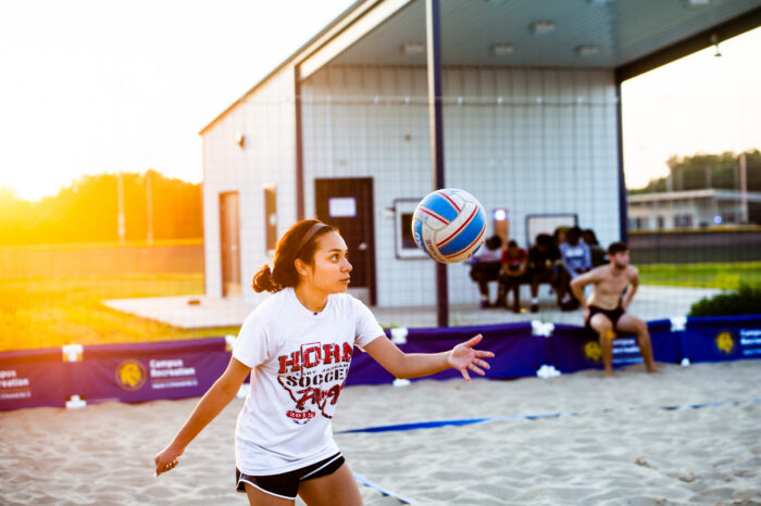 Girl standing in the sand, throwing a volleyball in the air.
