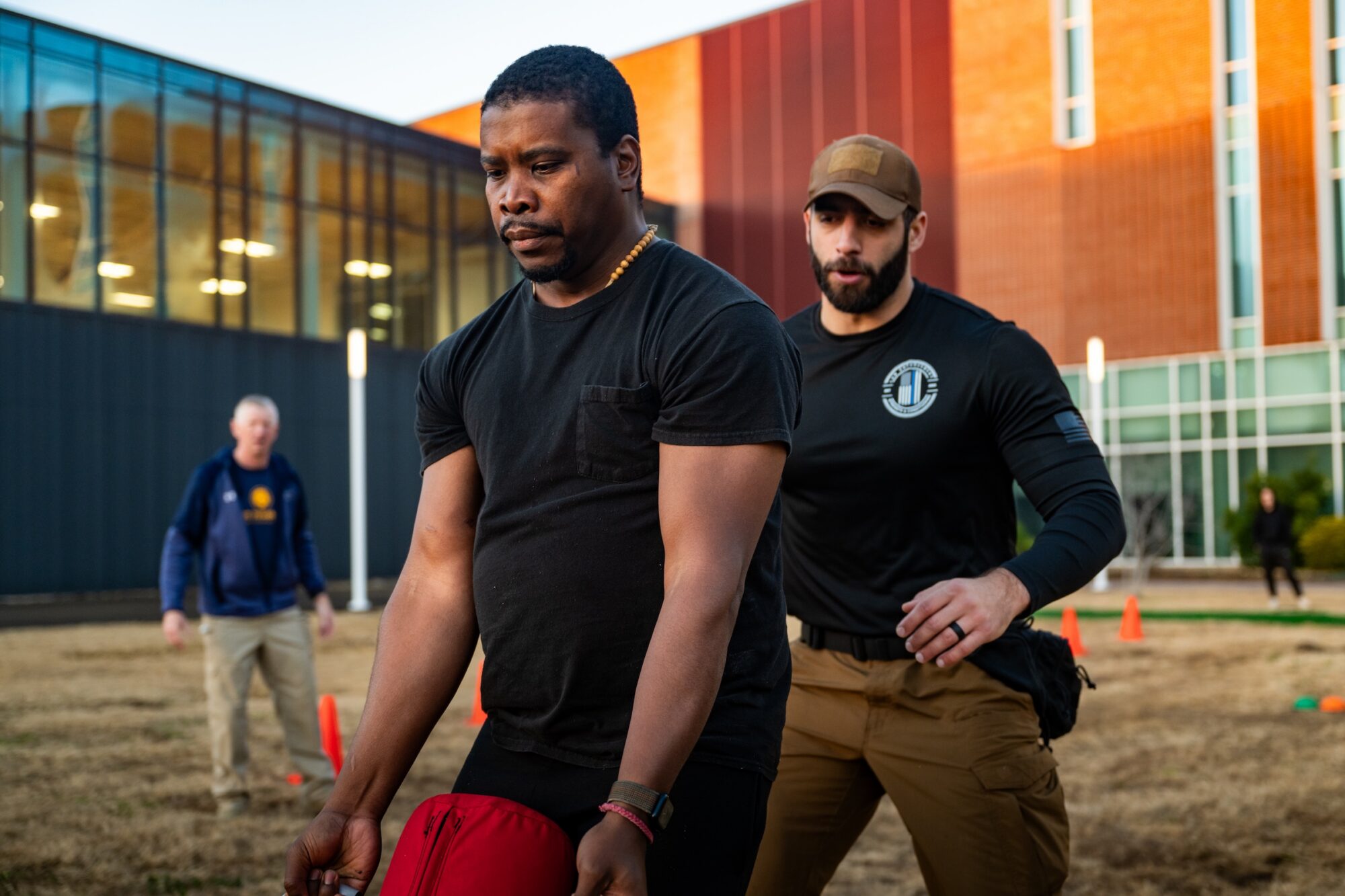 Person lifts a heavy red bag. Trainer is behind the person, with hand lifted to support the person if needed.