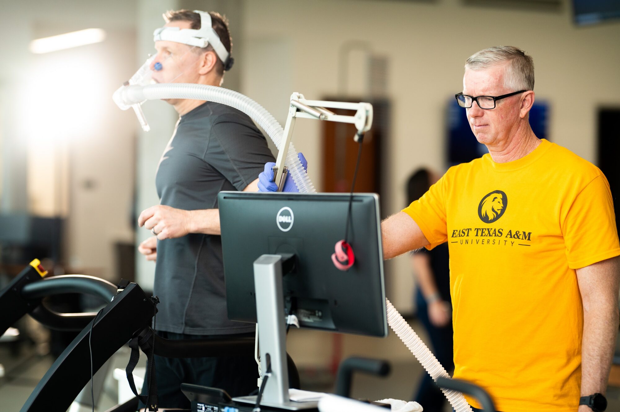 Person on the left is on a treadmill, hooked up to a hose device that runs from his nose and mouth to a computer. Employee on the right is monitoring data on a computer screen.