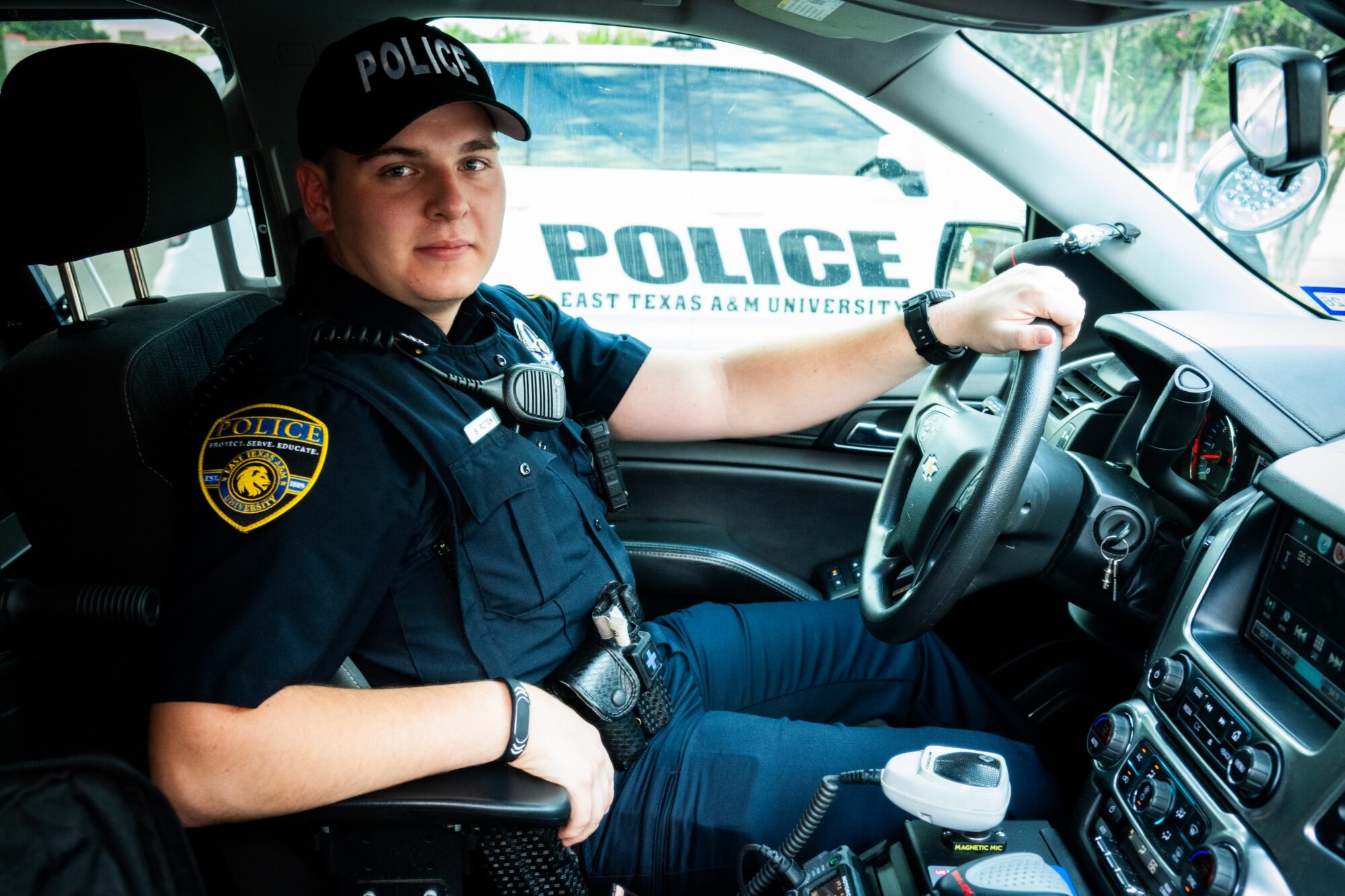 Police officer sits behind the wheel of a police car. His left hand is on the wheel and his right hand is on the arm rest. He is facing the camera and smiling slightly.