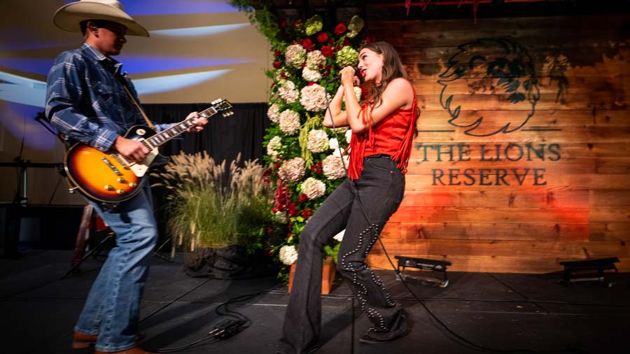 Person on left is playing guitar with cowboy hat on, blue wester shirt and jeans with books. Person on left is singing into a microphone, wearing a red shirt with black western jeans. The two people are facing each other.