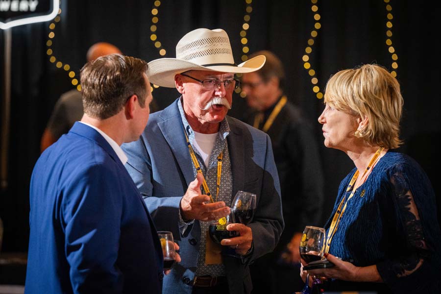 Man in cowboy hat talks with two people who are on either side of him, listening. They each hold a glass of wine in their hand.