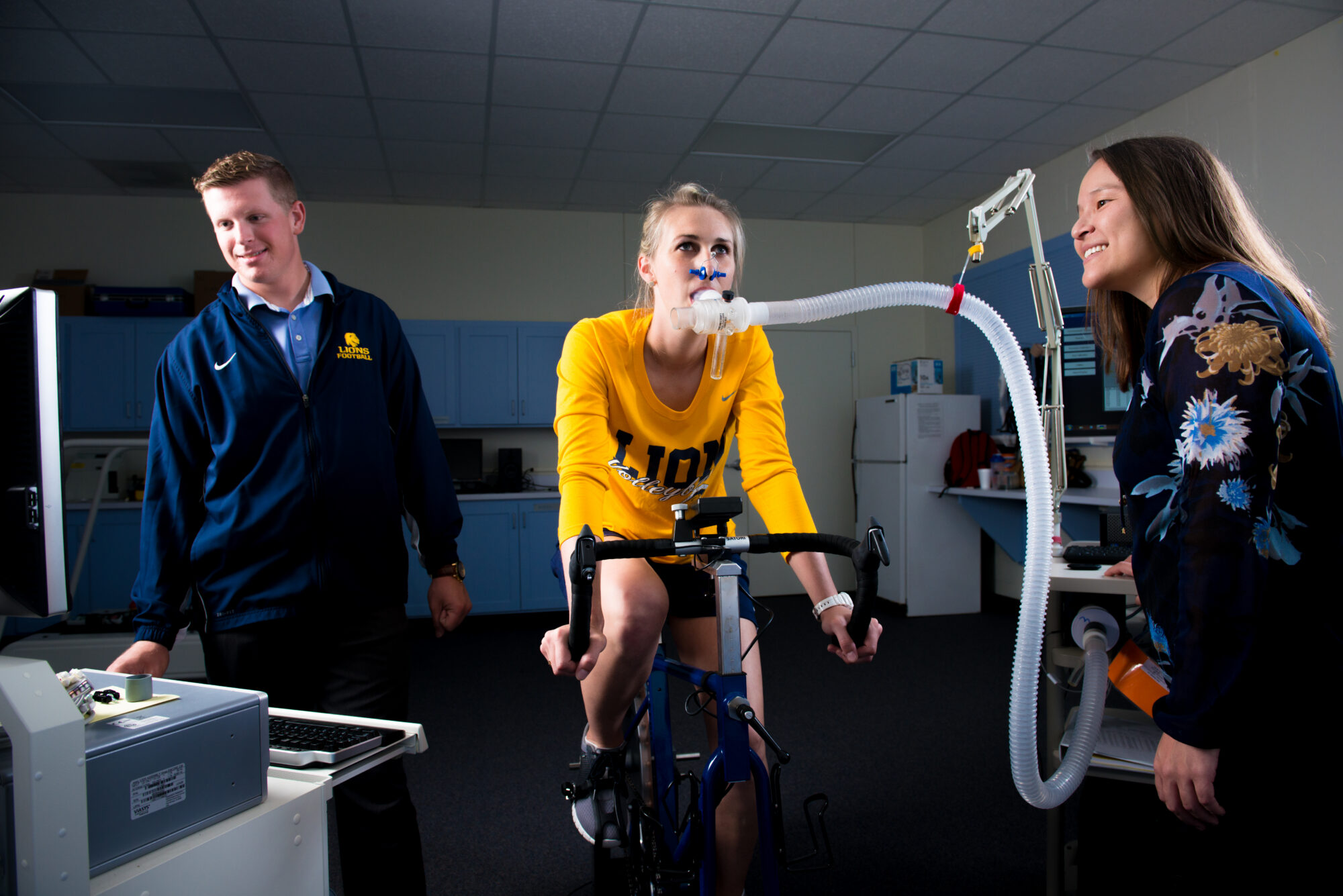 A woman riding on a stationary bike as two people watch.