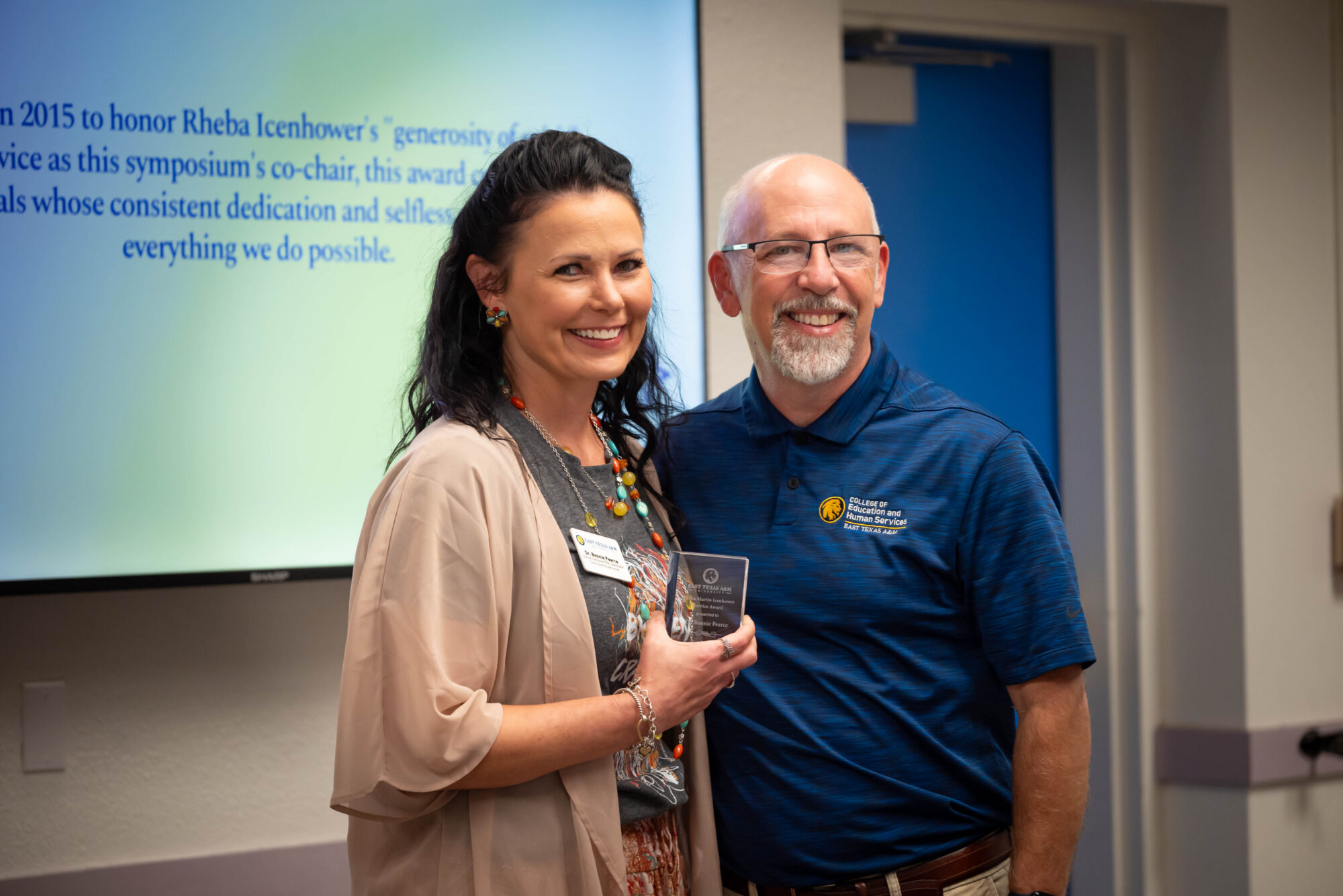 A man and a woman standing together and smiling, the woman holding a small glass trophy.