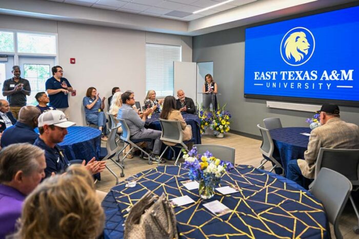 A large room is shown with a blue screen at the front of the room. The screen has the university's logo. A speaker at a podium is on the left side of the screen. Several people sit at tables and stand along the back wall.