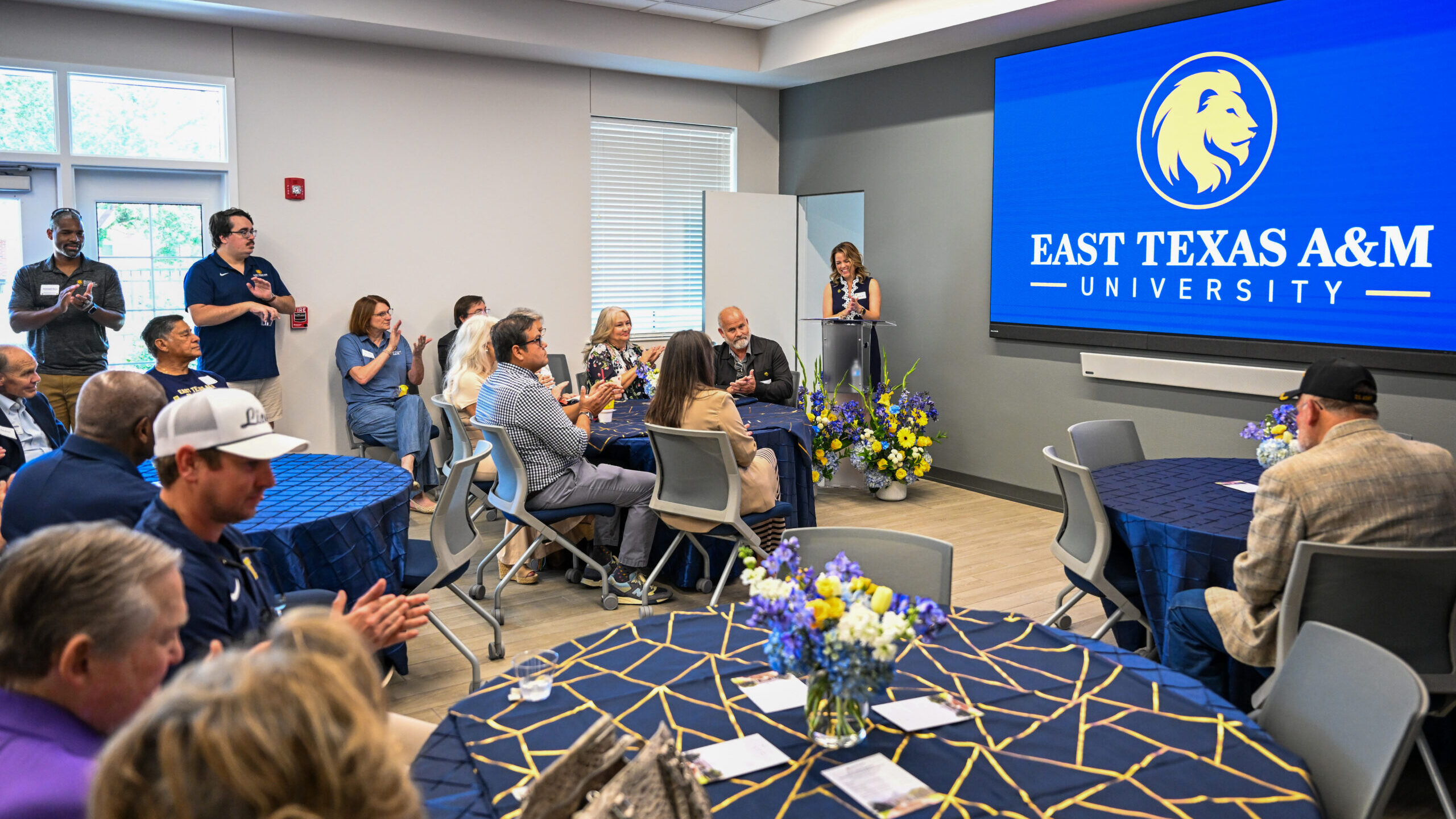 A large room is shown with a blue screen at the front of the room. The screen has the university's logo. A speaker at a podium is on the left side of the screen. Several people sit at tables and stand along the back wall.
