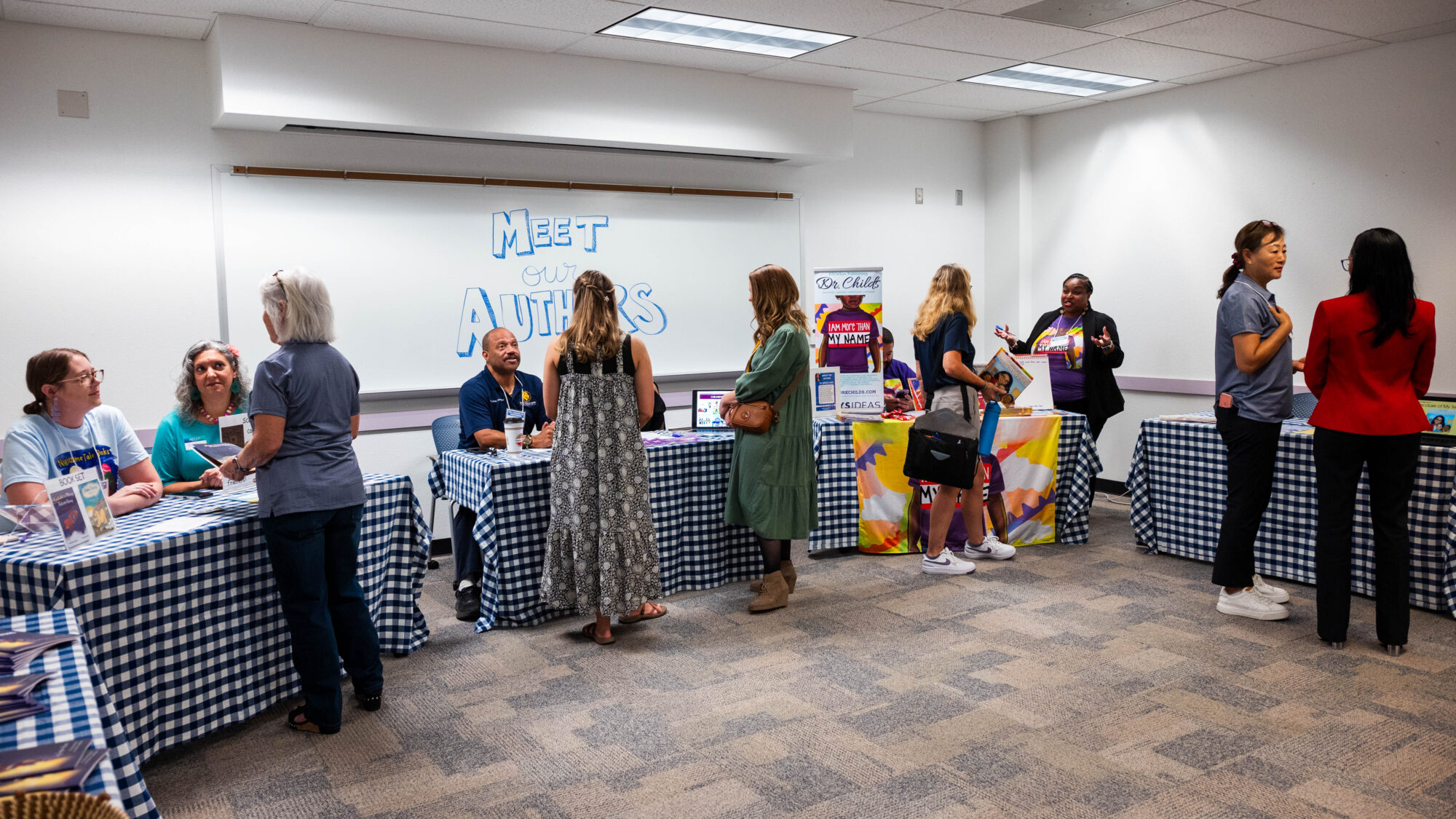 Several people sitting behind tables and several people walking around the tables, with a sign reading "Meet the Authors" in the background.