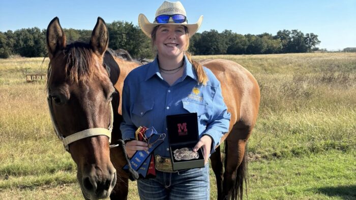 A person in western attire posing with a decorated belt buckle next to a brown horse.