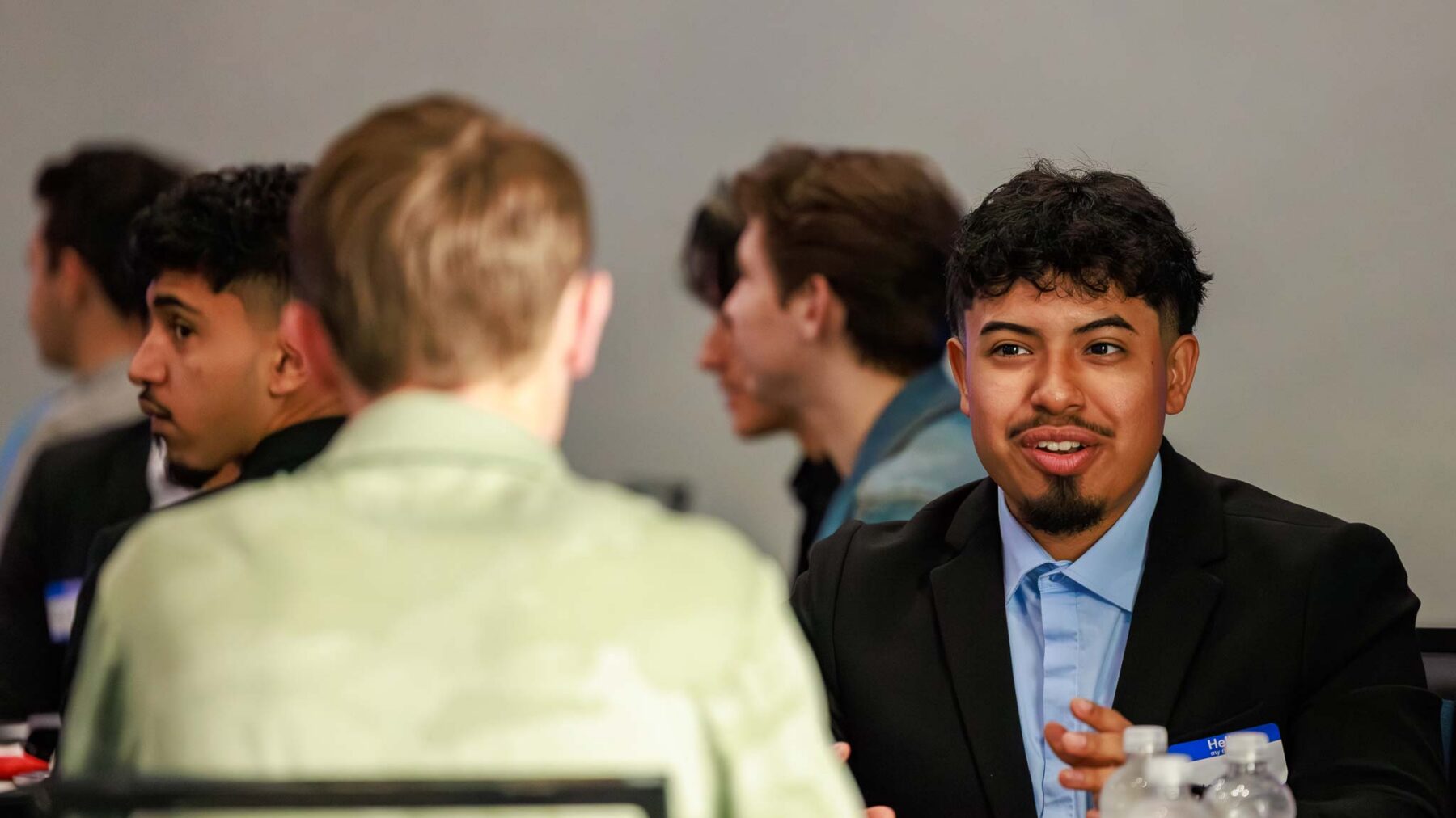 A young man seated at a table talks with another young man.