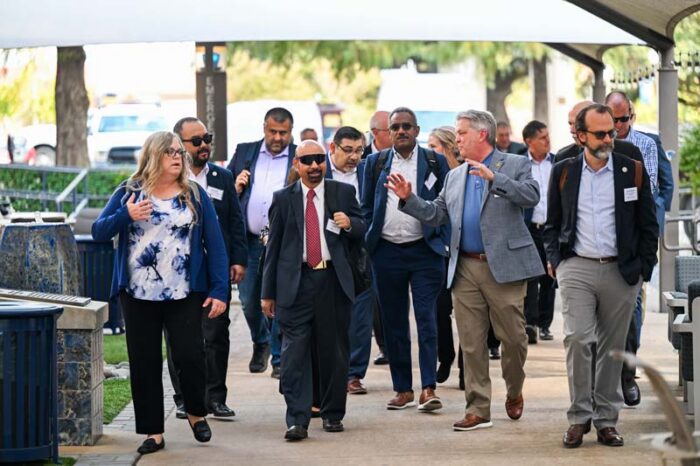 Several people walk down the sidewalk, toward the camera. Some are talking. A man toward the front is talking to someone on his right and gesturing with his hands.