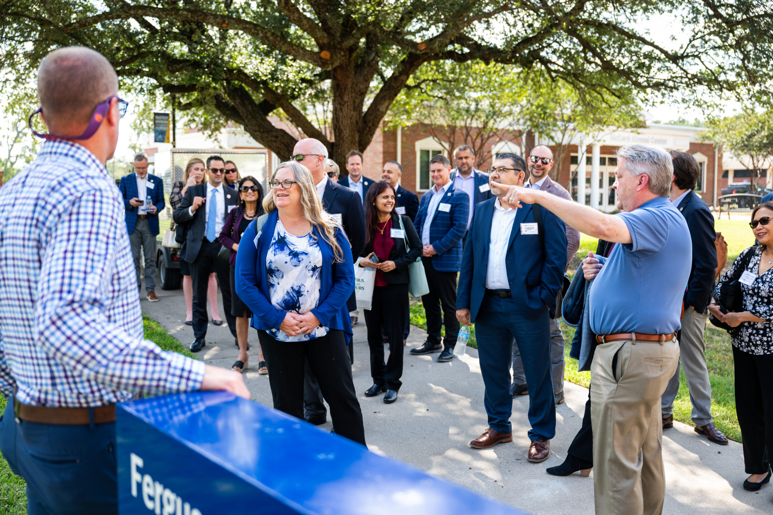 A man points to a building while several people standing beside him on the sidewalk look toward where he is pointing. Many are smiling, and many are wearing sunglasses.