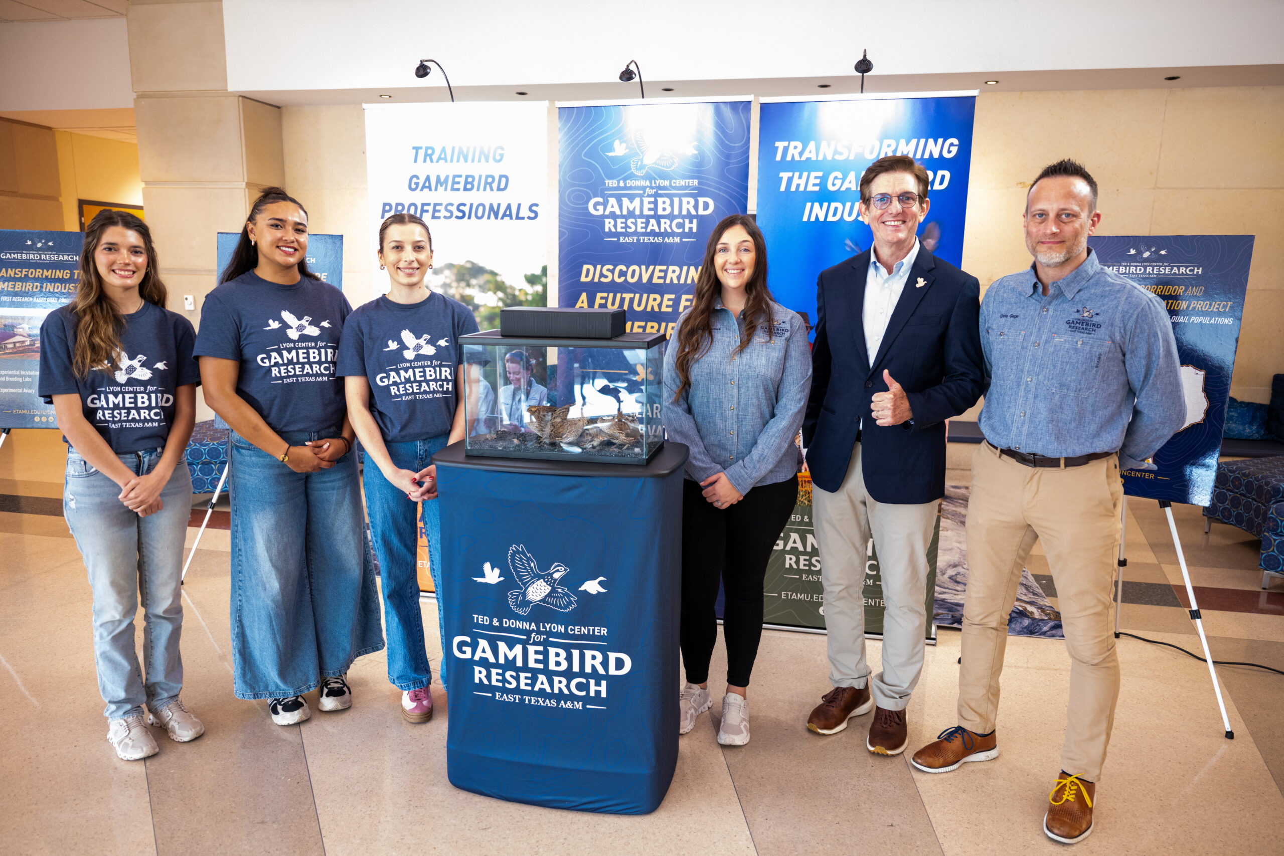 Three people stand on each side of a podium that is holding a glass case with quail inside.