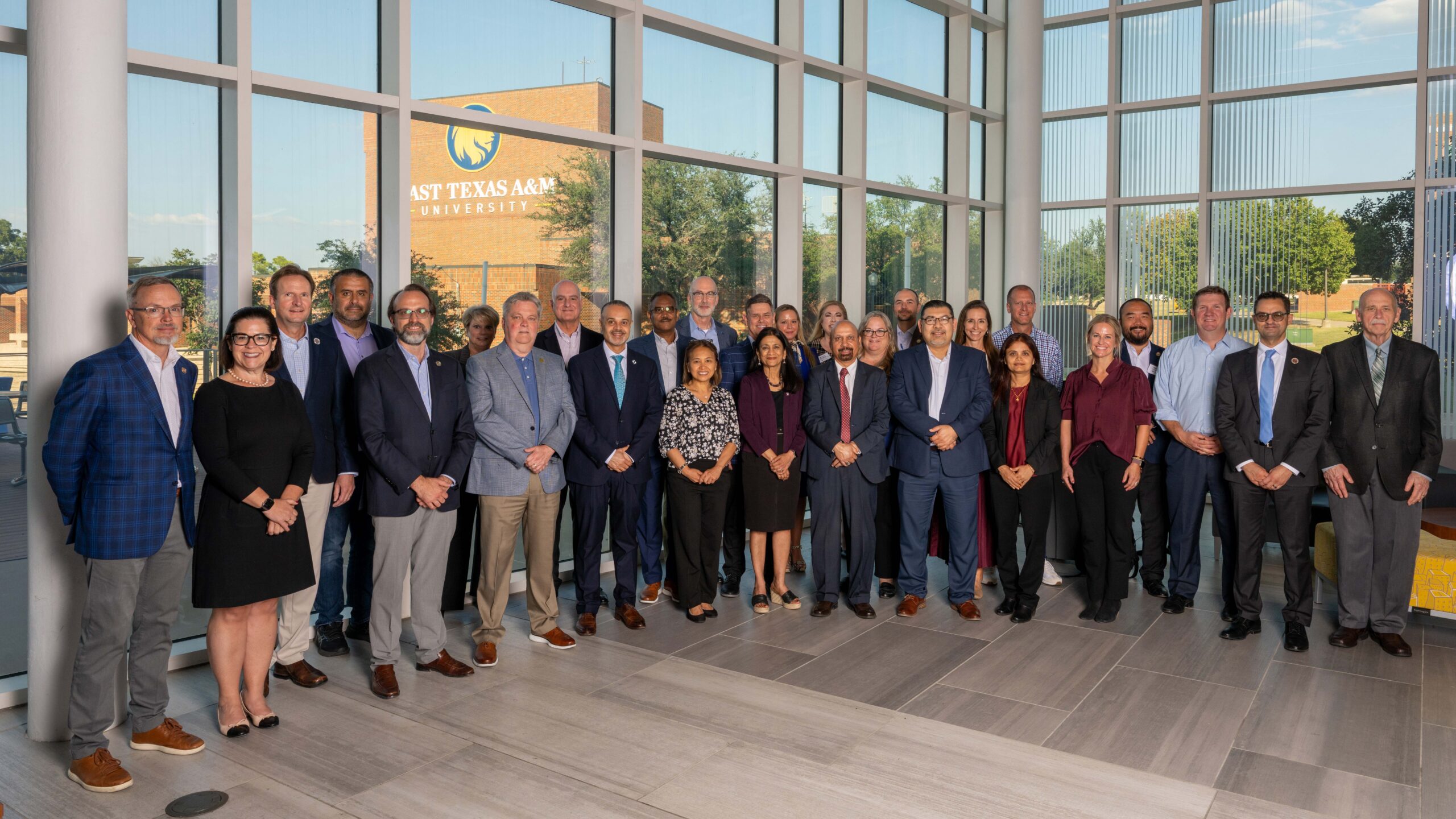 A group of around 40 people stand in front of a glass wall and smile for the camera.