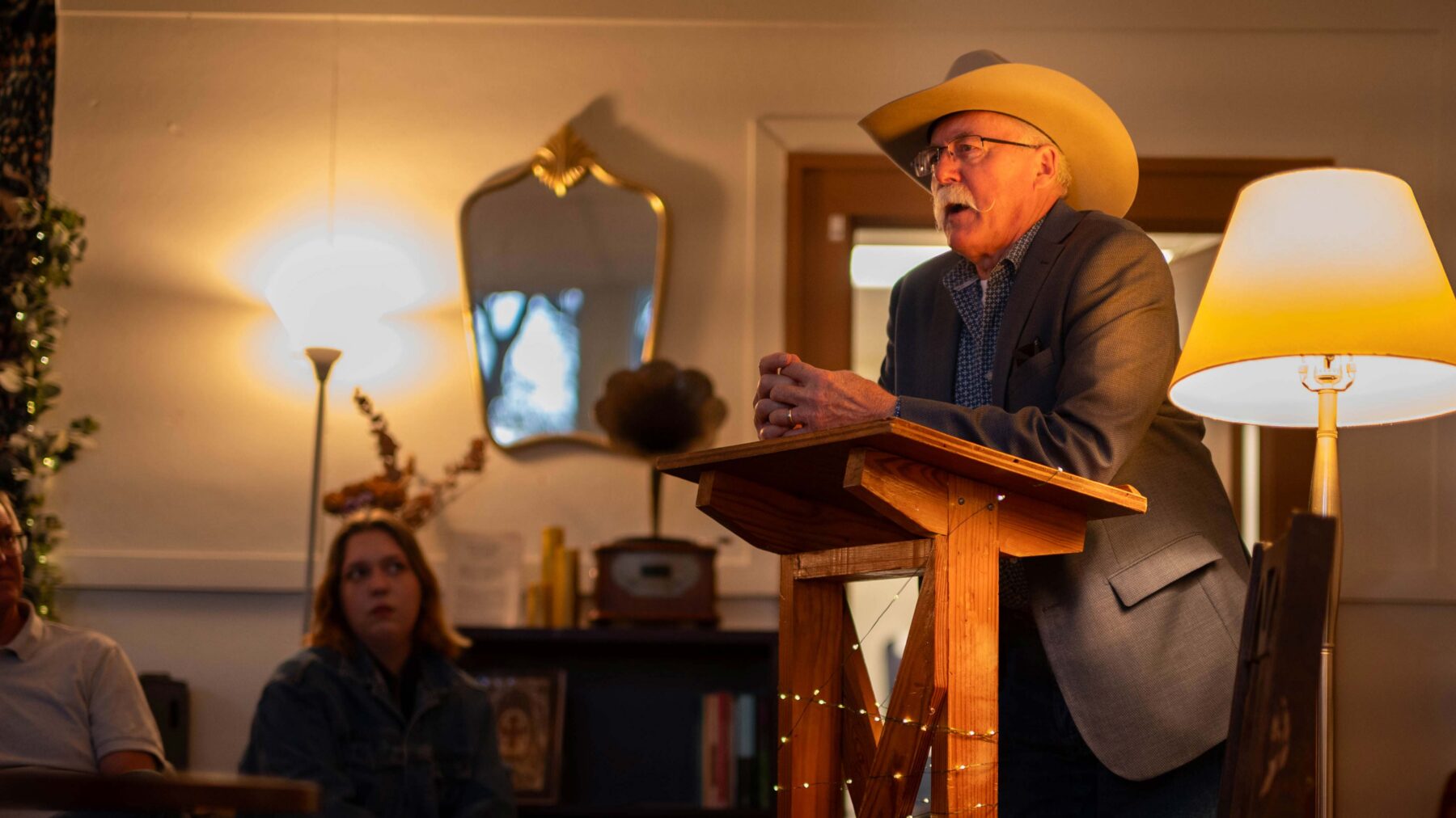 A man wearing a cowboy hat stands at a podium talking, while a woman in the background looks on.