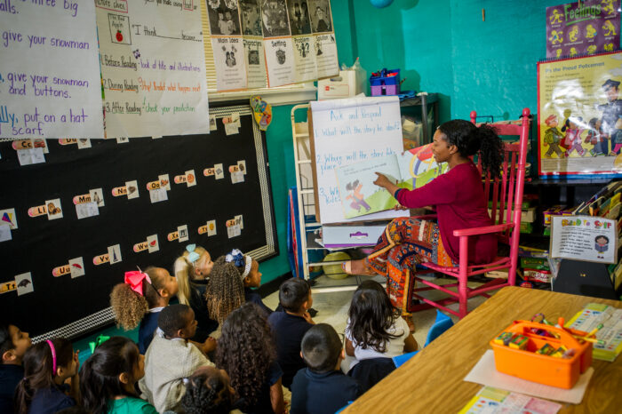 A woman teaching in a classroom with children sitting on the floor.