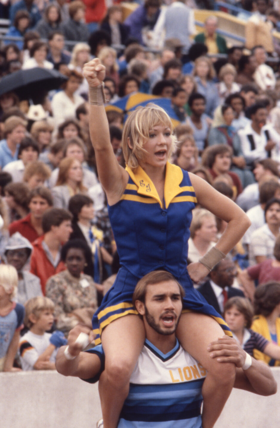 Two cheerleaders at a football game, with one sitting on the other's shoulders.