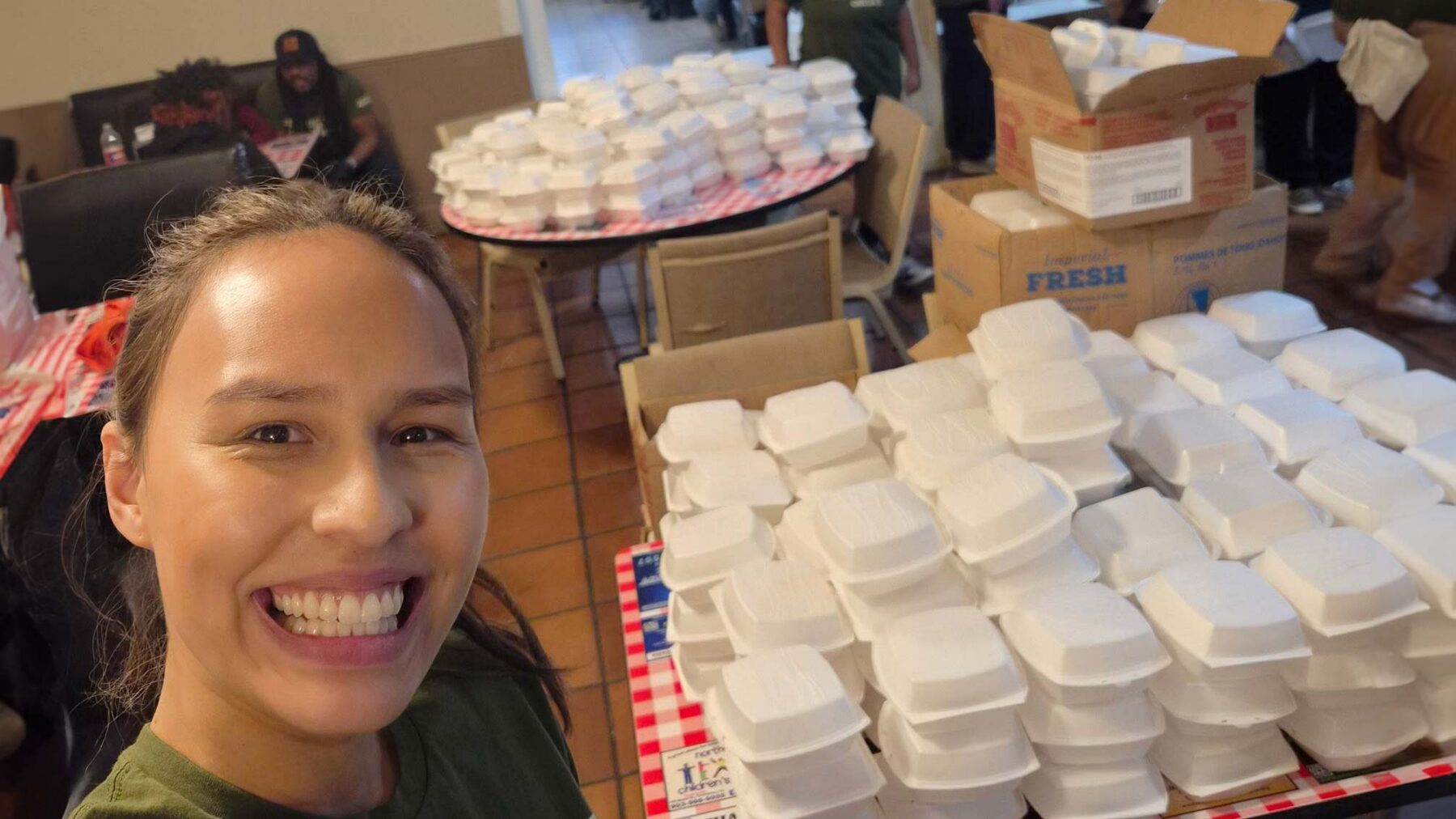 Two tables are piled with white styrofoam containers, presumably filled with food. On the bottom left of the frame, a woman smiles at the camera.