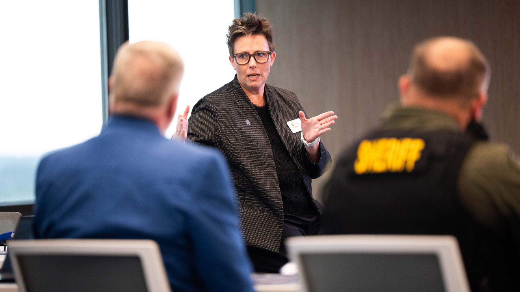 A woman is standing and talking in front of two men seated in chairs.