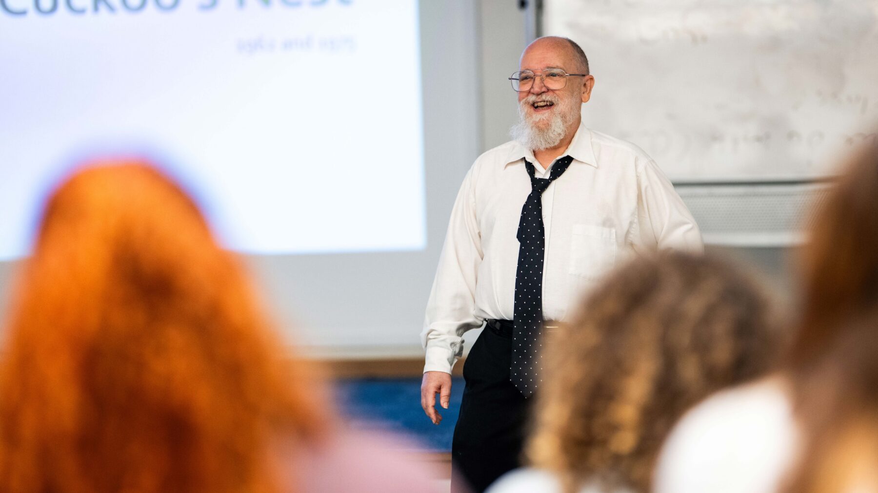 Man wearing a tie smiles, while standing in front of a class of students.