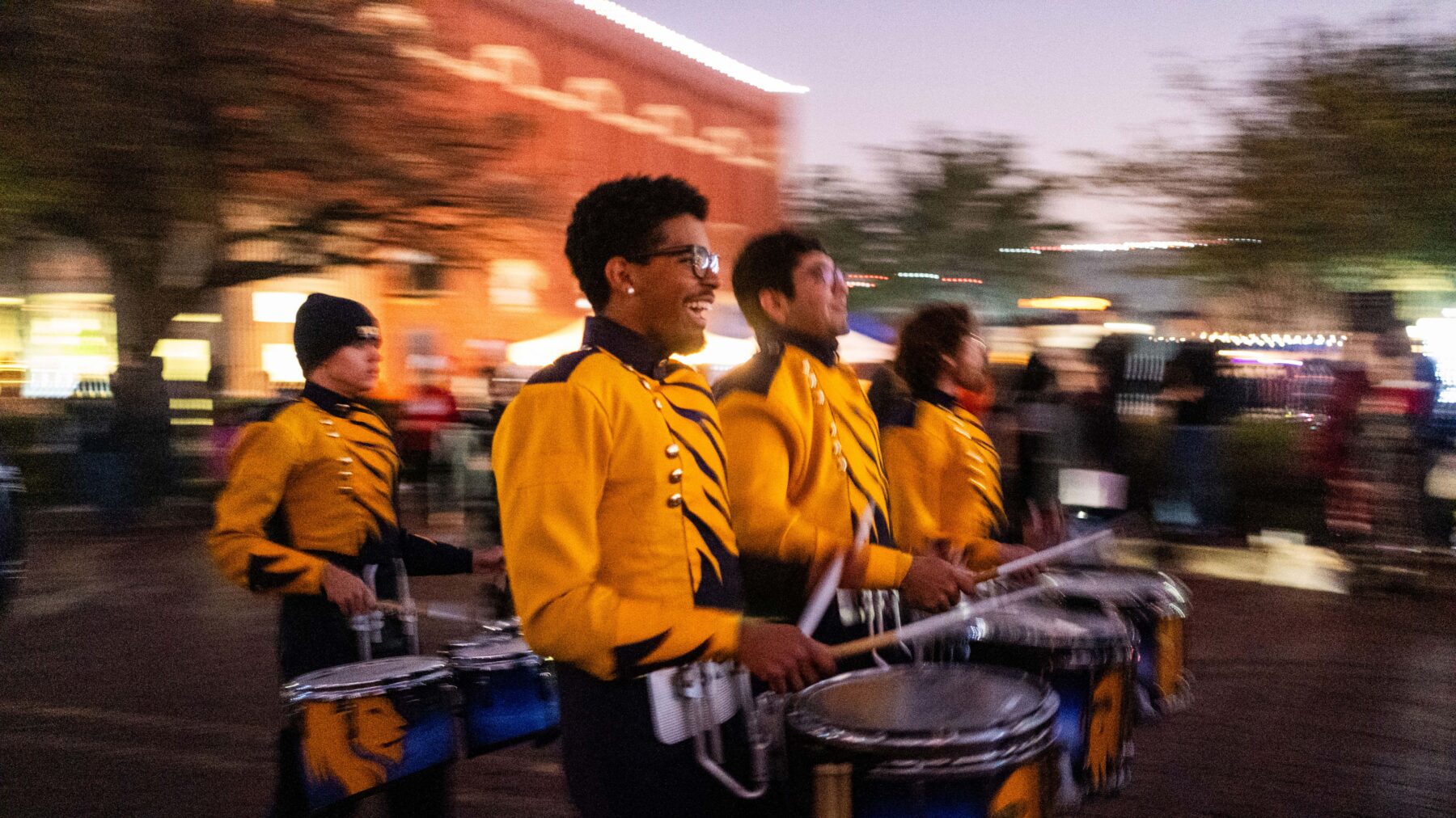 Members of the marchiing band pass by the camera wearing gold uniforms on top and black on the bottom. They are carrying blue and gold drums with the gold Lion head on them.
