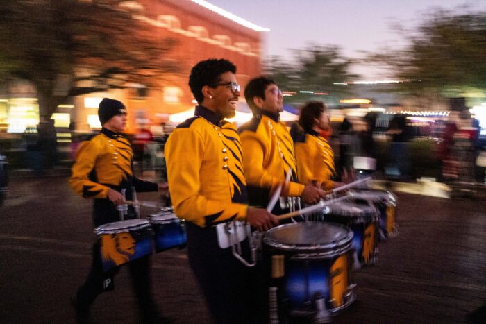 Members of the marchiing band pass by the camera wearing gold uniforms on top and black on the bottom. They are carrying blue and gold drums with the gold Lion head on them.