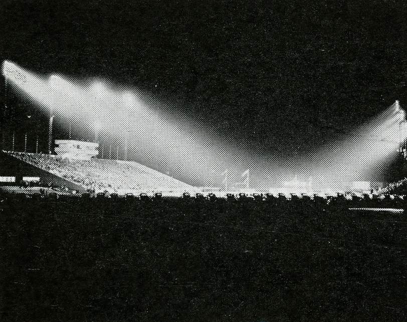 A black and white photo of a football stadium during a game at night.