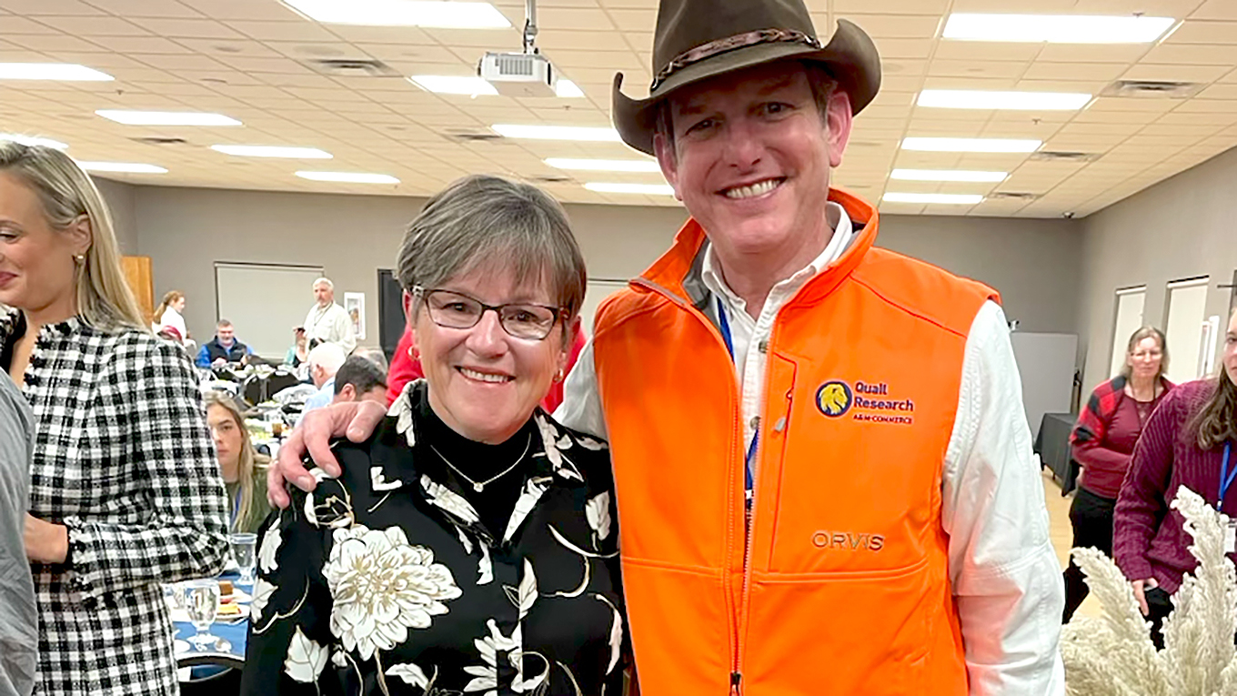 A man stands to the right wearing a brown Stetson hat, white button-up, and Blaze orange hunting-style vest. His arm is on the shoulder of a woman to his right with short-cropped hair wearing a black top with white and gold lotus flowers