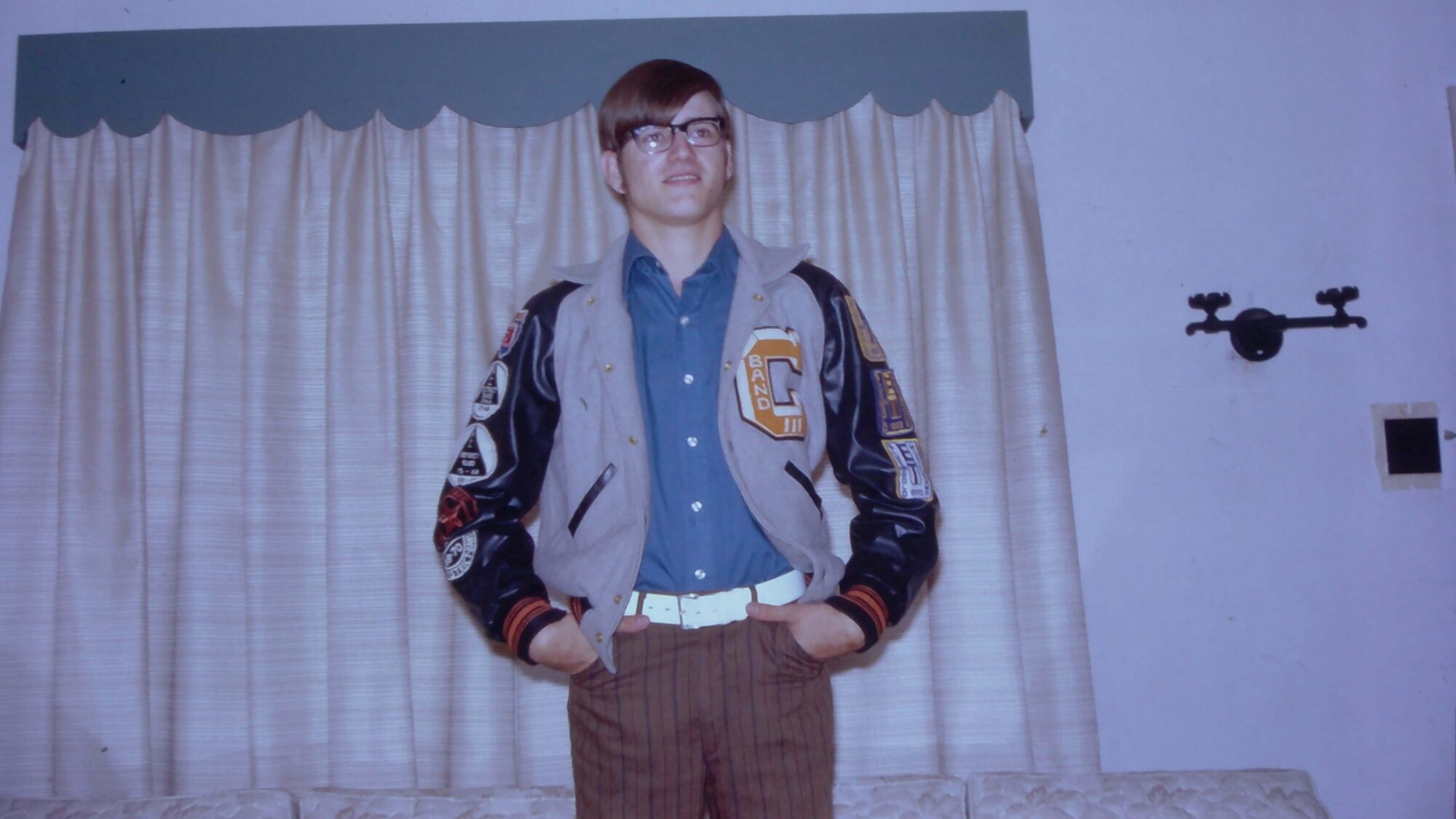 A teenager faces the camera wearing a high school letter jacket that says "Band" on the front. He has his hands in his pockets and is wearing a white belt and rimmed glasses.