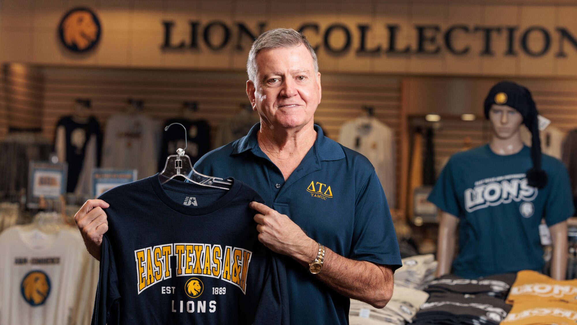 A man smiles at the camera while holding a t-shirt on a hanger from within a store. Clothing merchandise, branded with East Texas A&M logo, surrounds him.