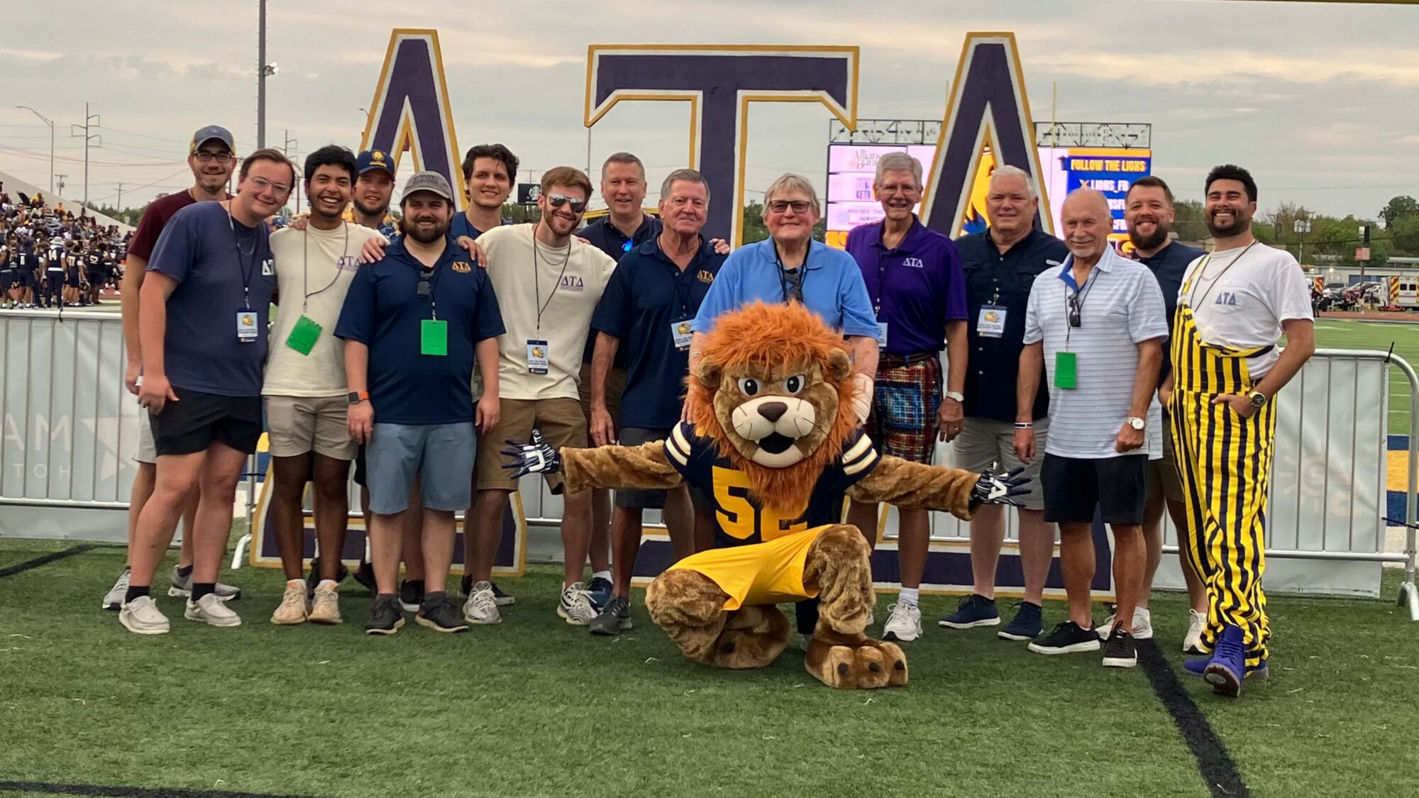 A group of around 15 people smiles for the camera as they stand on a football field. A lion mascot (costume) sits in front of them with his arms outstretched.