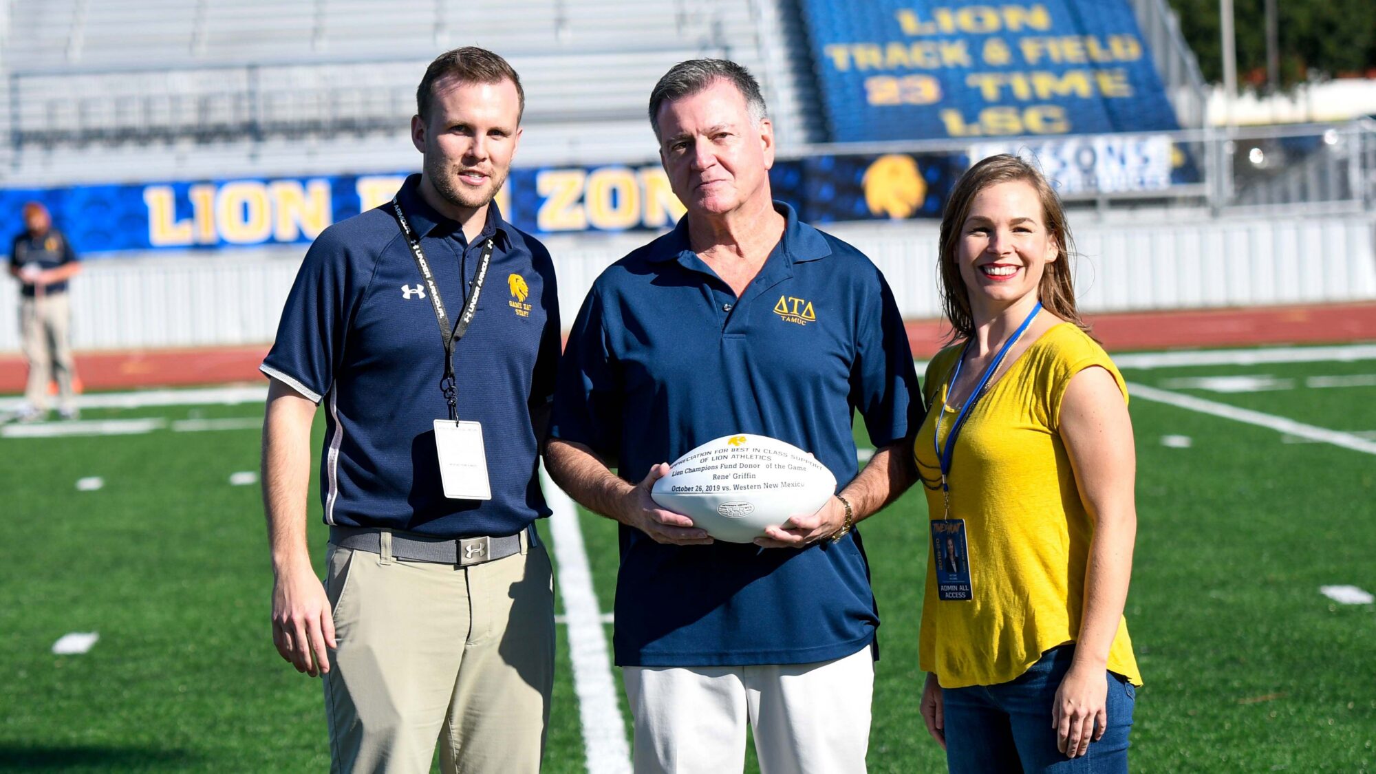 Three people smile for the camera as they stand on a football field. the man in the middle is holding a white football.